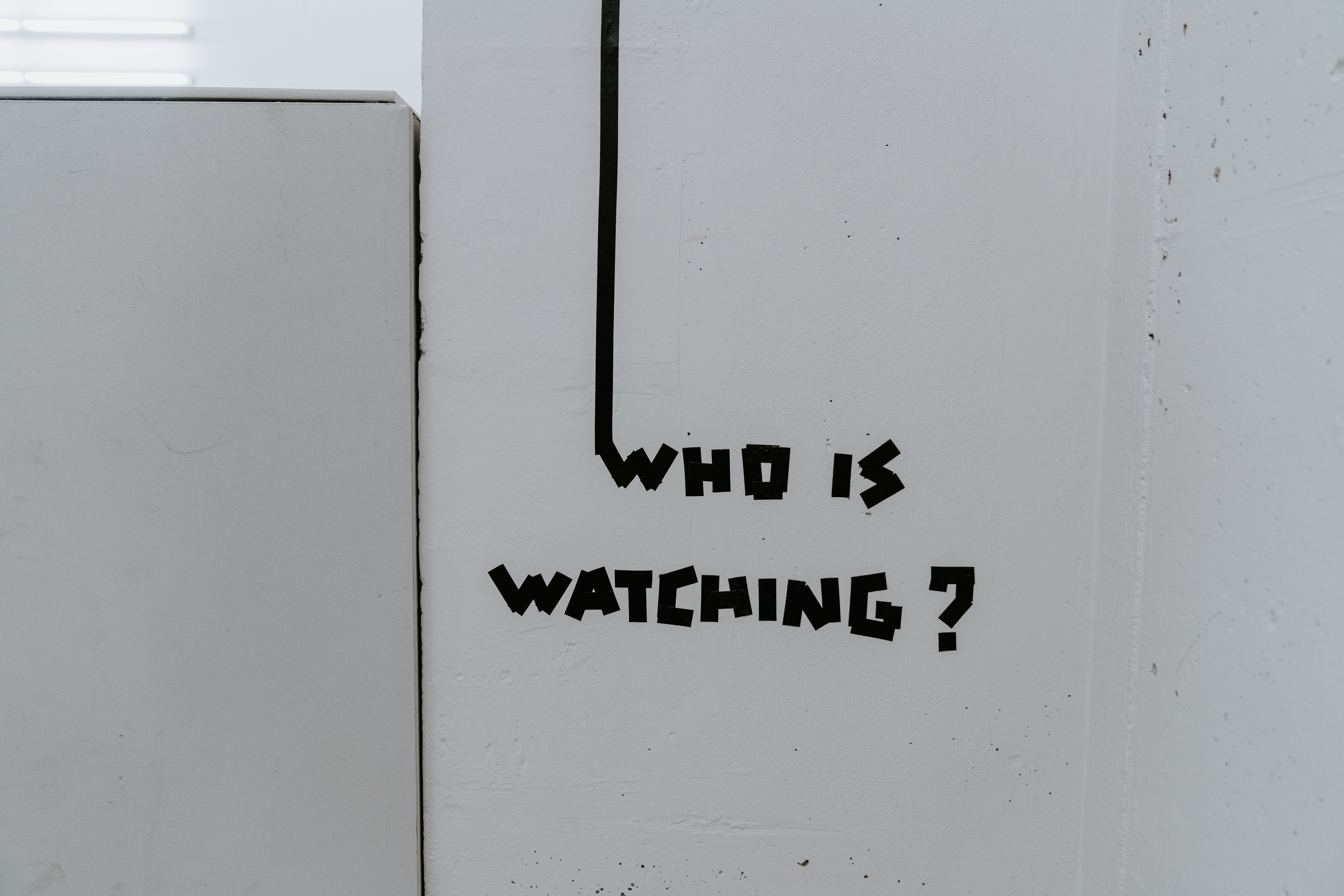 Tag mural who is watching ? en noir sur mur blanc.