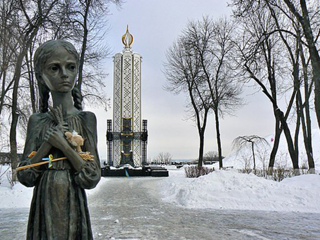 Photographie extérieure en format paysage et en couleurs du Mémorial aux victimes du Holodomor. Au premier plan, sur la gauche, apparaît une statue de fillette ayant le regard dans le vide et portant des épis de blé contre sa poitrine. Au fond, on voit le mémorial blanc et or, de forme étroite et haute. Les arbres sont dénués de feuilles et le sol est recouvert de neige. La lumière est une lumière blanche et froide d'hiver.