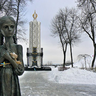 Photographie extérieure en format paysage et en couleurs du Mémorial aux victimes du Holodomor. Au premier plan, sur la gauche, apparaît une statue de fillette ayant le regard dans le vide et portant des épis de blé contre sa poitrine. Au fond, on voit le mémorial blanc et or, de forme étroite et haute. Les arbres sont dénués de feuilles et le sol est recouvert de neige. La lumière est une lumière blanche et froide d'hiver.
