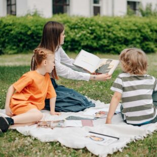 Femme qui lit un livre à deux enfants assis dans l'herbe.