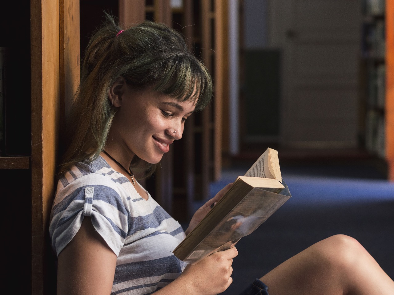 Une adolescente lit un livre assise par terre dans une bibliothèque.