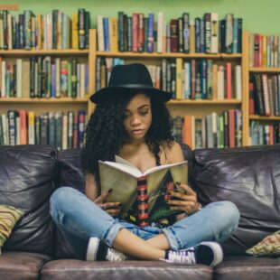 Femme assise sur un canapé en train de lire avec une bibliothèque en fond.