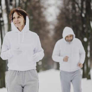Photographie en couleurs de format paysage montrant une femme au premier plan et un homme au second plan dehors dans un paysage de neige et d'arbres. Les 2 personnes sont en train de faire leur footing. L'ambiance est hivernale avec une lumière très blanche.