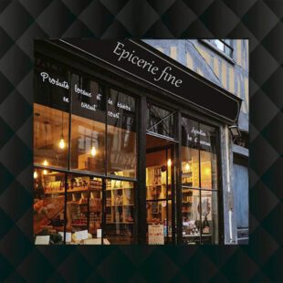 Photographie en couleurs encadrée par un fond noir ayant l'aspect capitonné. La photographie en extérieur montre une épicerie éclairée à l'intérieur et on peut voir les etagères à travers la vitre. Sa devanture est noire et porte le nom d'épicerie fine. Elle est mitoyenne à une maison à colombages gris/bleu