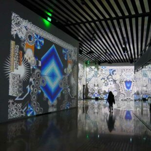 Photographie intérieure en paysage montrant une silhouette parcourant une installation immersive d'art numérique. La hauteur sous plafond est haute et la personne parait petite. Sur tous les murs sont projetés des dessins géométriques. L'ambiance est à la fois sombre et lumineuse