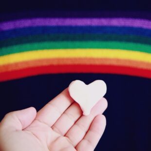 Une paume de main avec un cœur en pâte à modelé posé dessus. Présence du drapeau LGBT.