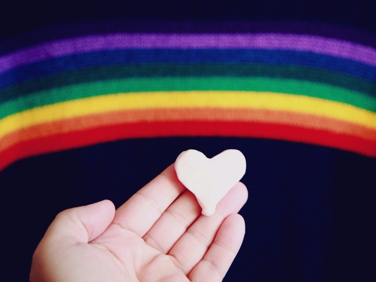 Une paume de main avec un cœur en pâte à modelé posé dessus. Présence du drapeau LGBT.